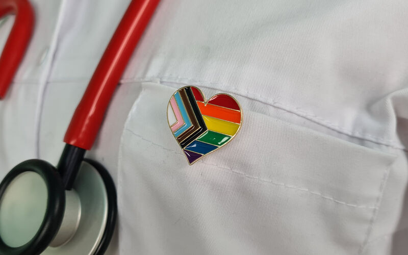 A doctor's coat displays a Progressive Pride pin in the shape of a heart.