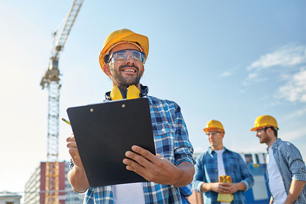 Construction safety inspector with clipboard and hard hat supervising site workers near a crane