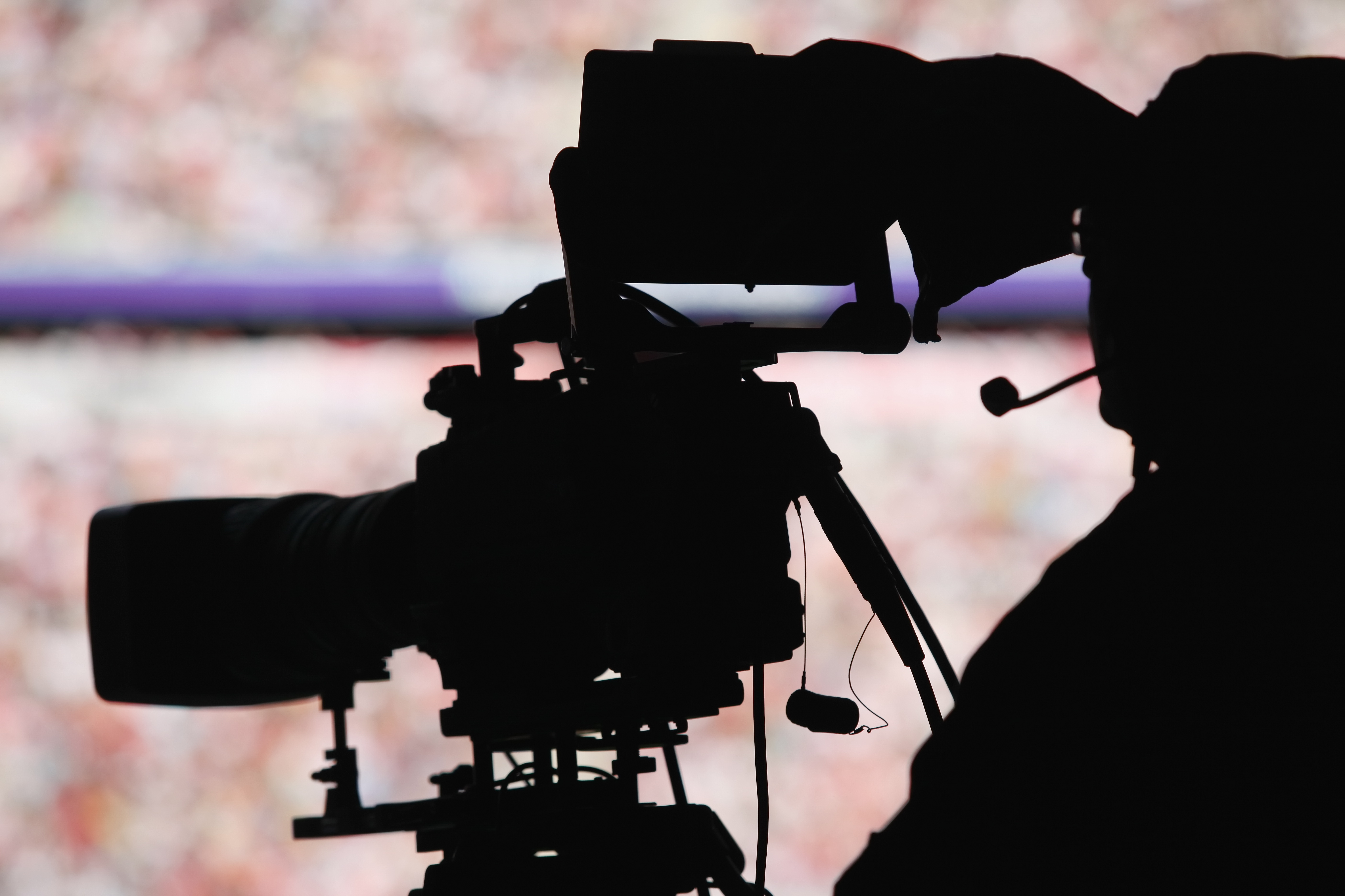 A cameraman uses AV production equipment during a sports broadcast, silhouetted in black against the crowd.