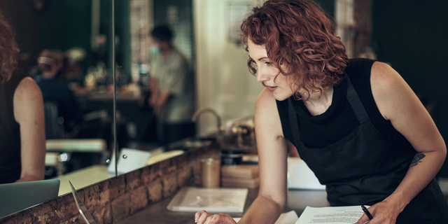 Shot of an attractive young hairdresser standing and using her laptop while calculating her finances in her salon