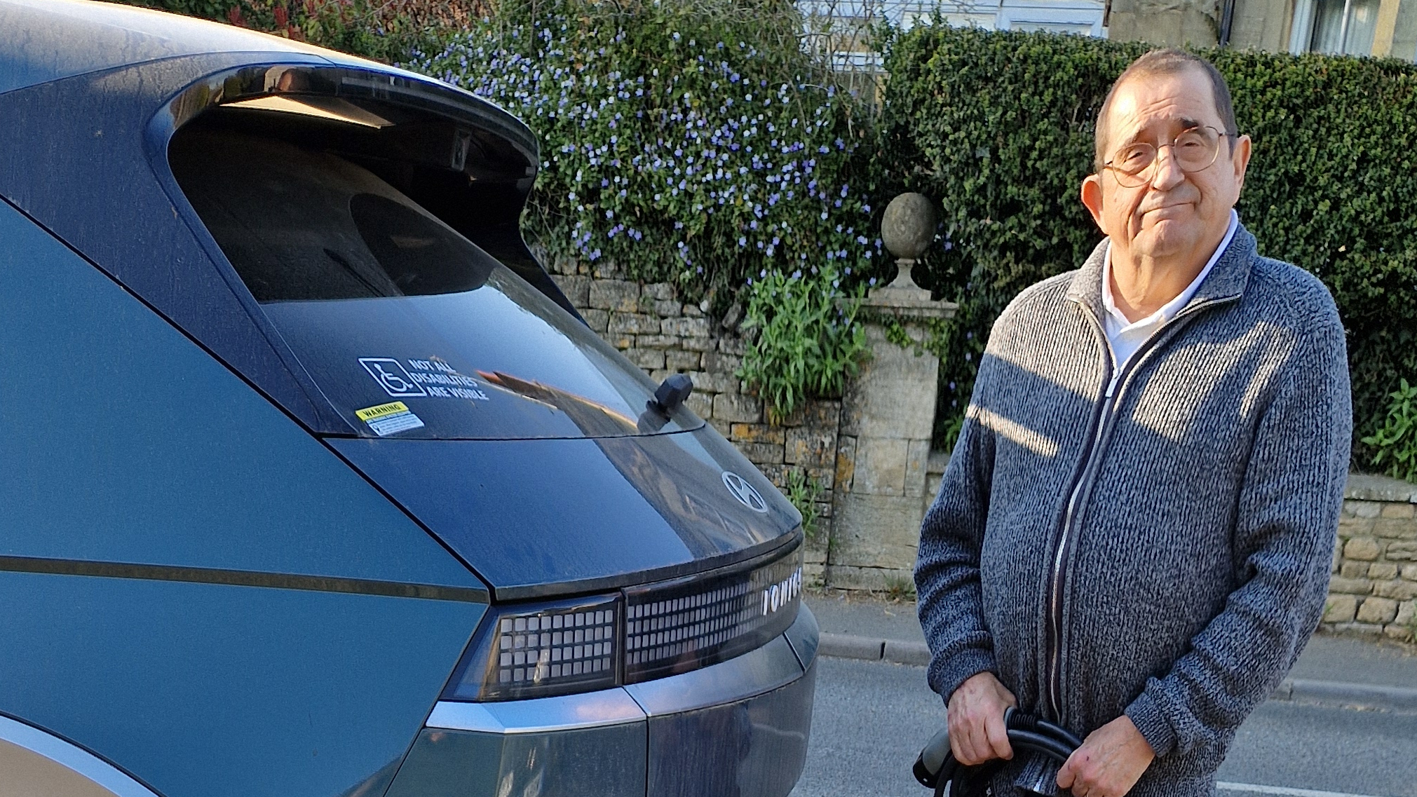 ChatGPT said: William Gee stands next to the rear of a blue Hyundai IONIQ 5, holding an EV charging cable in his hands. A disability parking permit is visible on the car’s rear window. He is wearing a grey zip-up sweater and glasses, with a gentle smile on his face. A stone wall and blooming flowers form the backdrop on a sunny day.