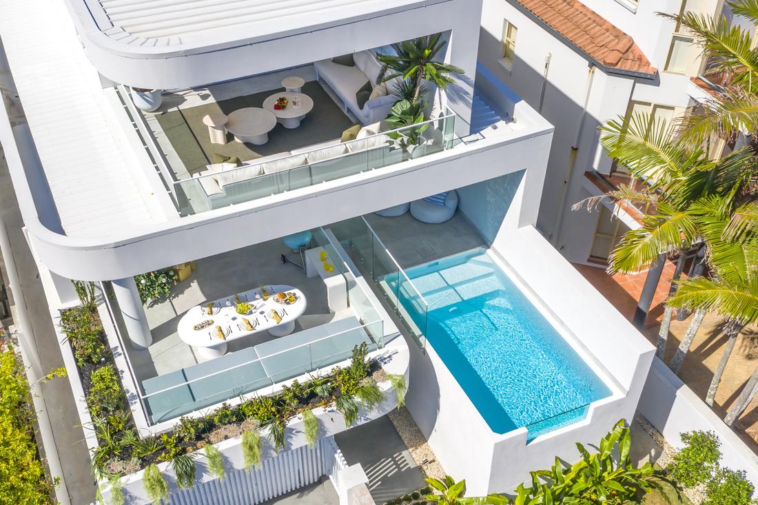 External aerial image of a new three-level tiered home. Second floor has a covered entertainment area with couch, table and plants. First floor has a raised pool to the right, and a covered entertaining area to the left with a table set for 6. There is a garden along the outer walls of the alfresco area with plants spilling over the edge.