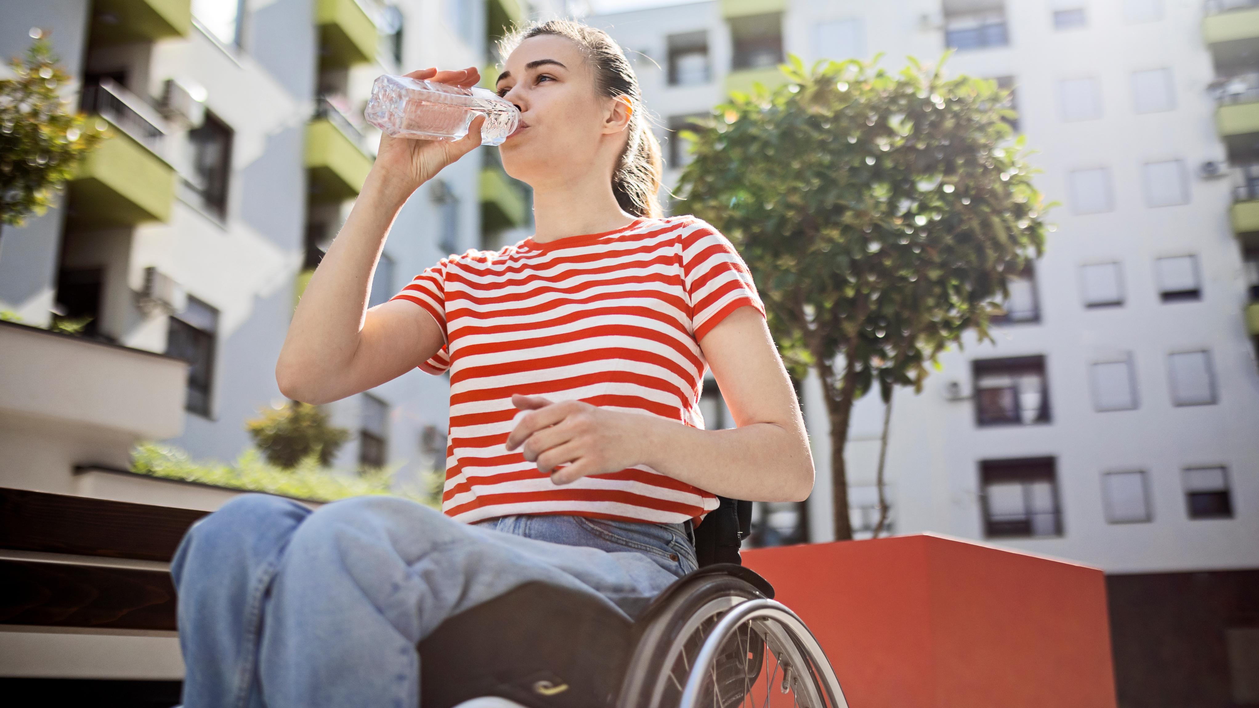 "A young woman in a wheelchair drinks water from a clear bottle while sitting outdoors. She wears a red and white striped t-shirt and blue jeans. The background features a modern apartment building with balconies, green plants, and a small tree nearby.