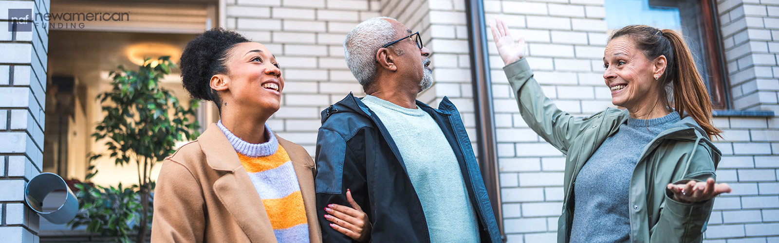 A young Black woman stands smiling arm-in-arm with an older Black man as they talk to a smiling white woman in front of a home