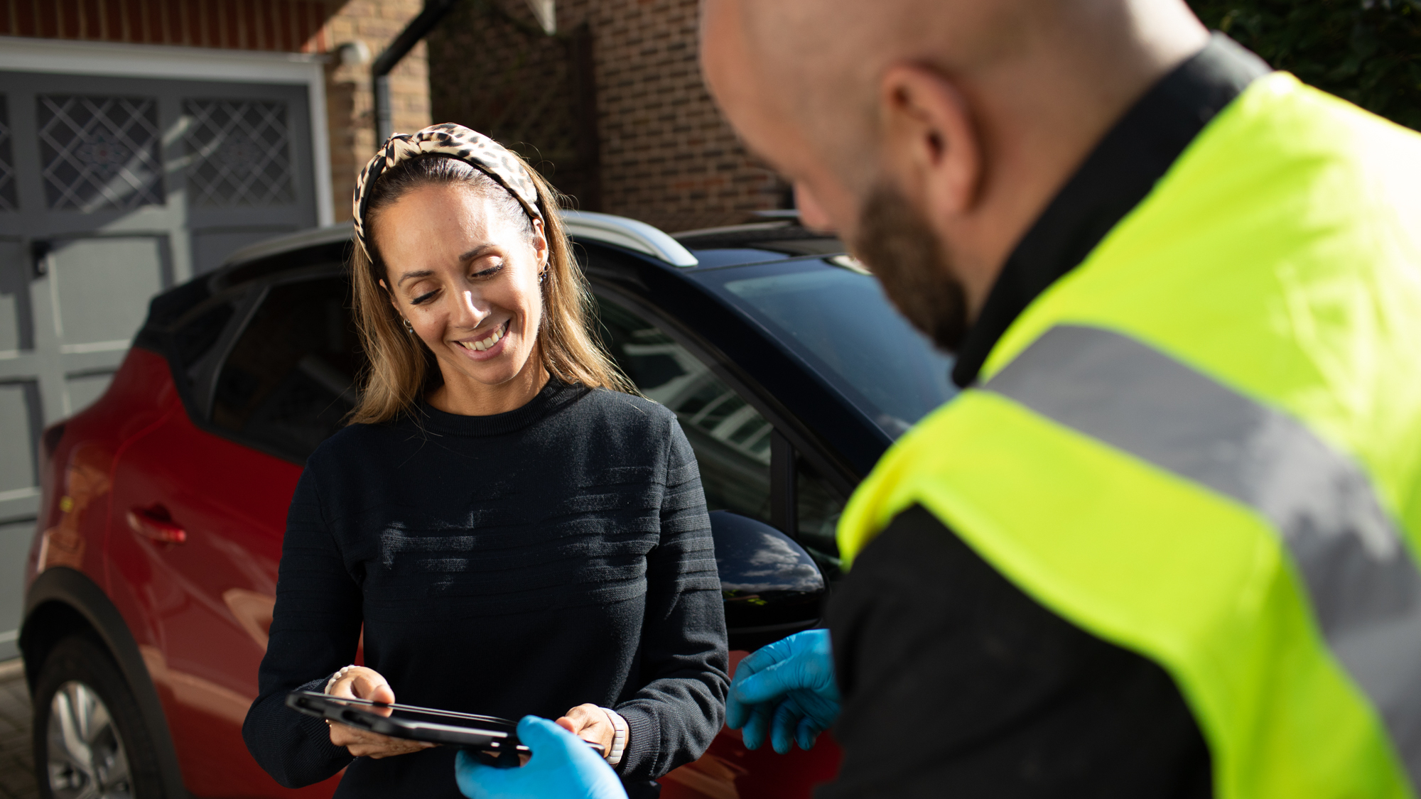 A smiling woman stands next to a red car, signing a digital tablet held by a man in a high-visibility vest and blue gloves. They appear to be outside a home, suggesting a customer receiving roadside assistance or a vehicle service.