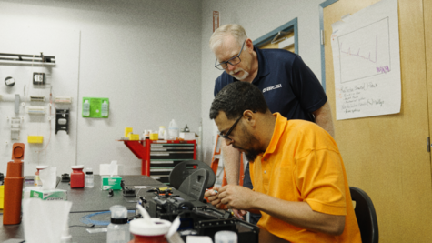 Instructor in a BICSI shirt overseeing a student performing precision fiber optic splicing at a workbench