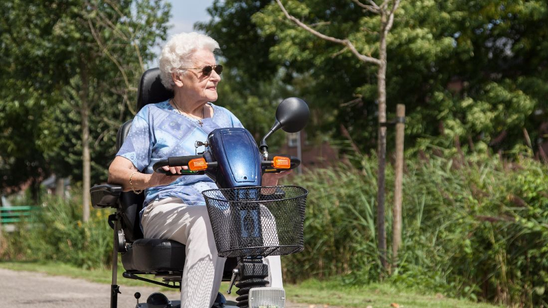 An older woman with white hair and sunglasses rides a blue mobility scooter along a paved path in a park. She wears a light blue patterned blouse and beige trousers. The background features green trees, grass, and a wooden fence, with sunlight casting a glow on the scene