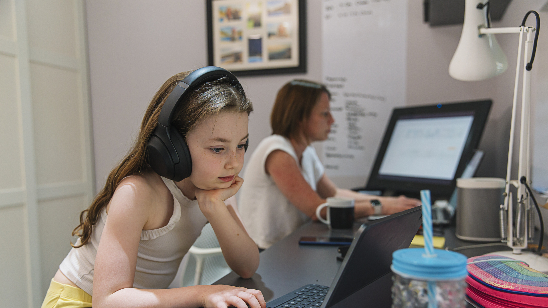 Young girl remote schooling in study. Mother is working in the background