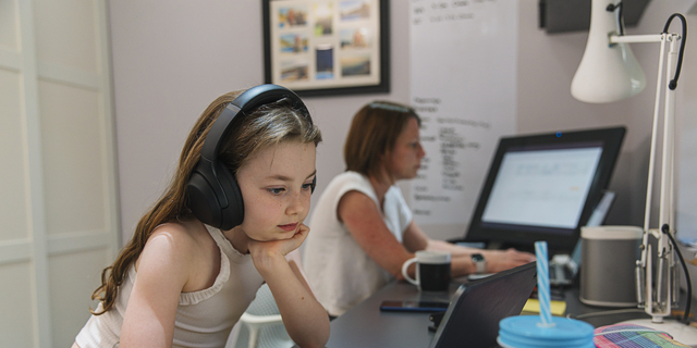 Young girl remote schooling in study. Mother is working in the background