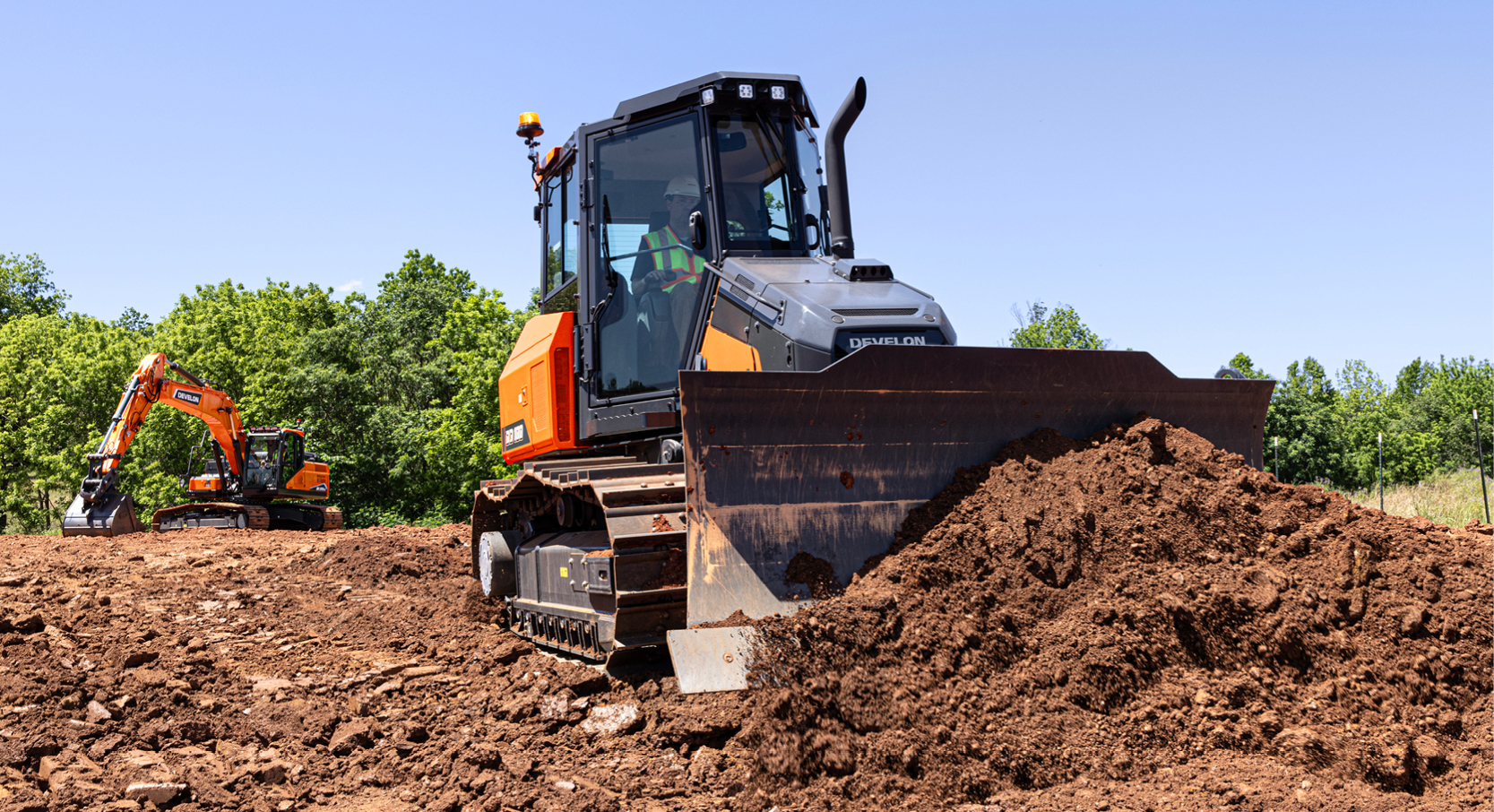 A DEVELON dozer grades dirt on a job site while a DEVELON excavator works in the background.