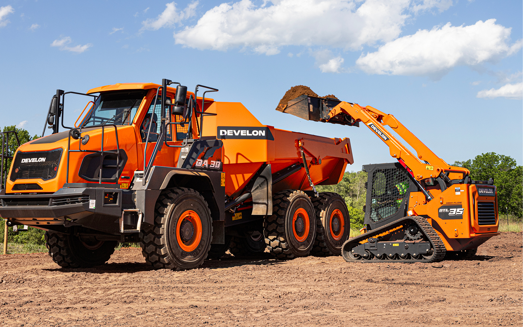 A DEVELON compact track loader prepares to empty a bucket of dirt into the bed of an articulated dump truck.