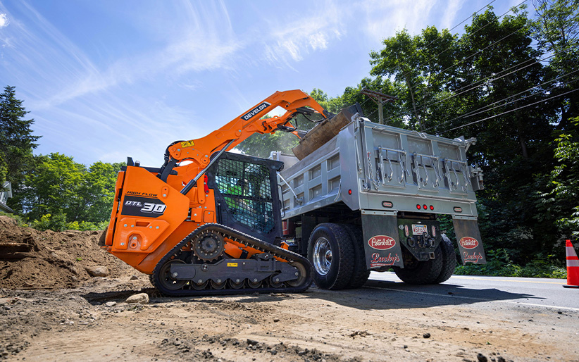 A DEVELON DTL30 compact track loader empties a bucket of dirt into a dump truck.