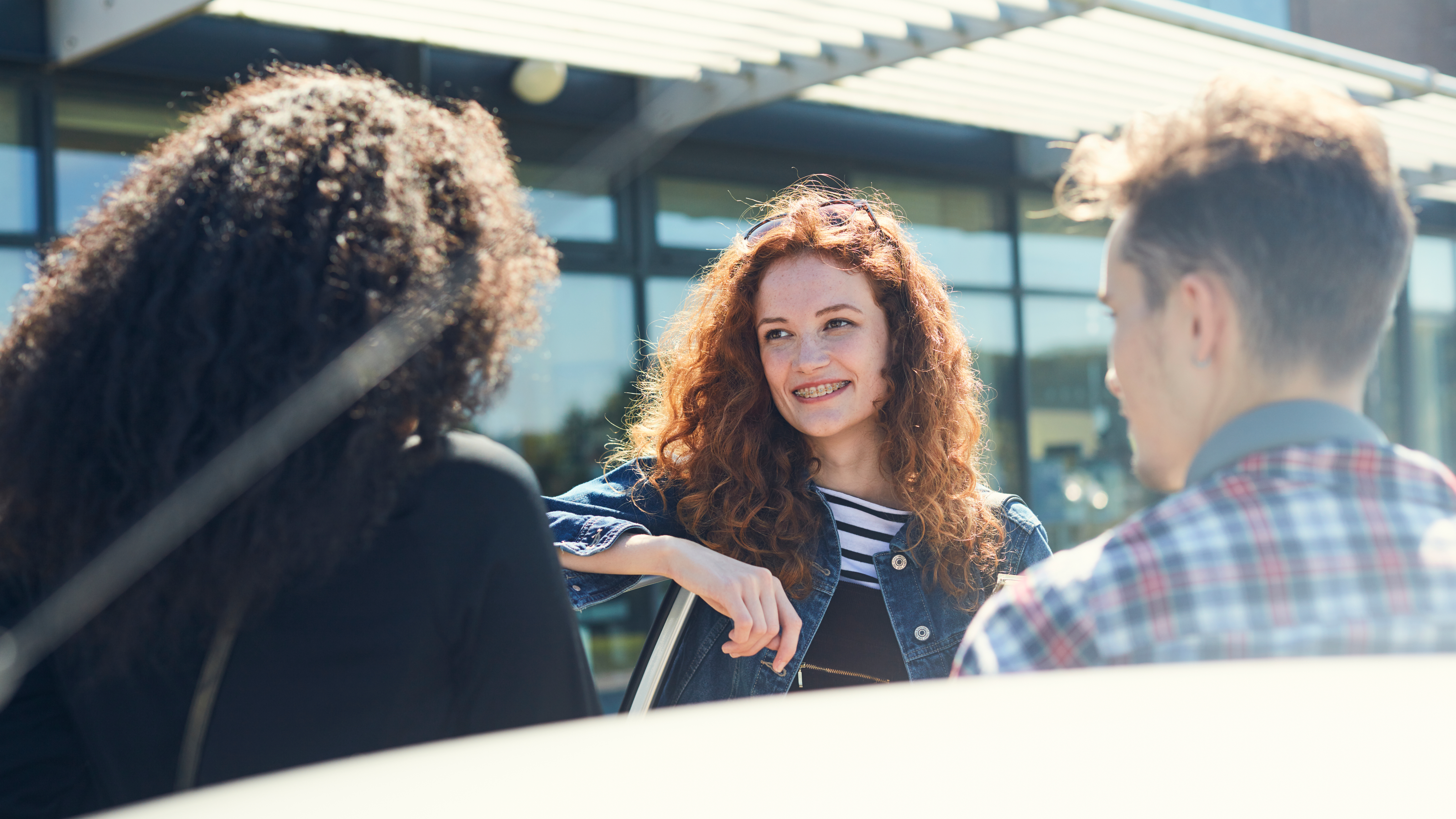 A young woman with curly red hair and braces smiles while talking with two other young people outdoors, standing in front of a modern building with large glass windows.