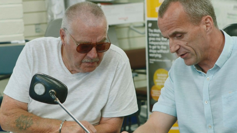 Two men sitting at a table, closely examining documents or a device. One man, wearing a white t-shirt and sunglasses, appears focused, while the other, in a light blue shirt, is explaining something. A mobility scooter mirror is visible in the foreground, and the background features posters and office equipment