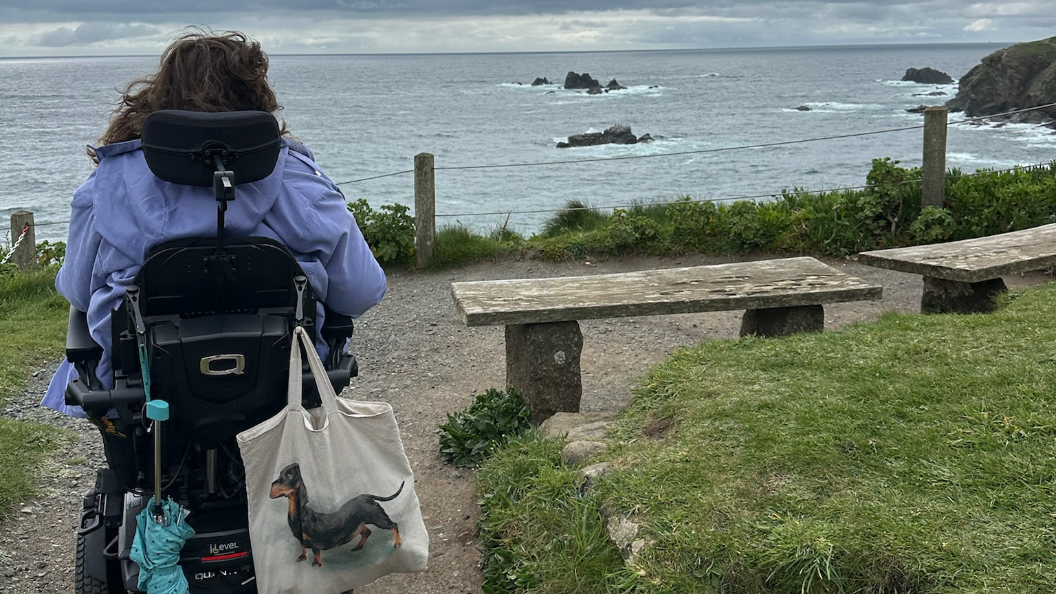Shona Louise is seen from behind, sitting in a power wheelchair on a coastal path. She faces the sea, with waves crashing on rocks in the distance, and carries a tote bag with a dachshund illustration on the back of her chair. The sky is cloudy, and stone benches line the path.