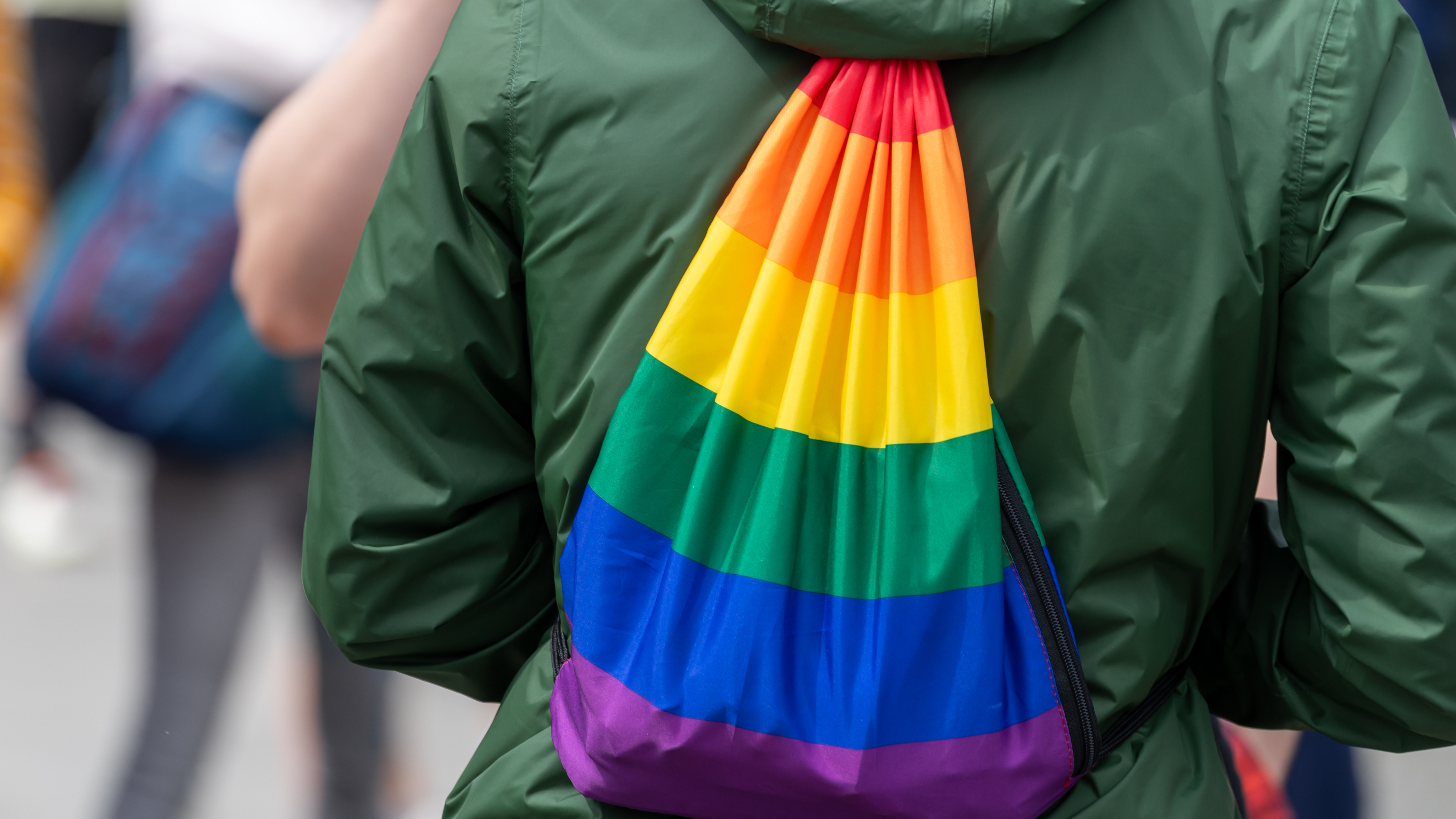 Close-up of a person wearing a green jacket and a drawstring backpack decorated with rainbow stripes. The image is taken from behind during a Pride event, with a crowd visible in the background.