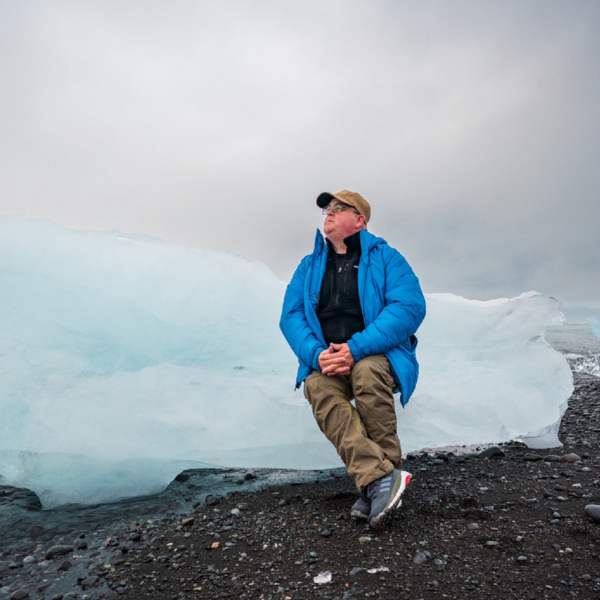 A man in a blue coat sits on a block of blue ice on the coast of Iceland