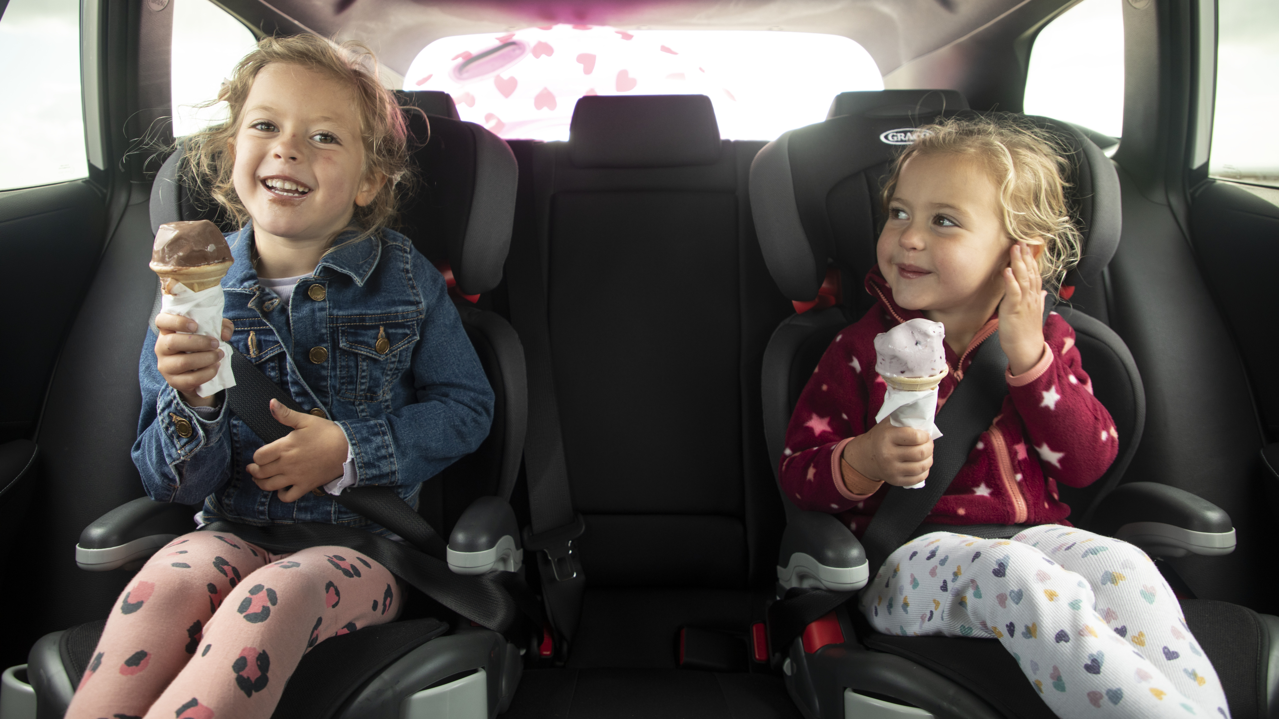 Two young girls sit in the back seat of a car, secured in car seats and smiling while eating ice cream cones.