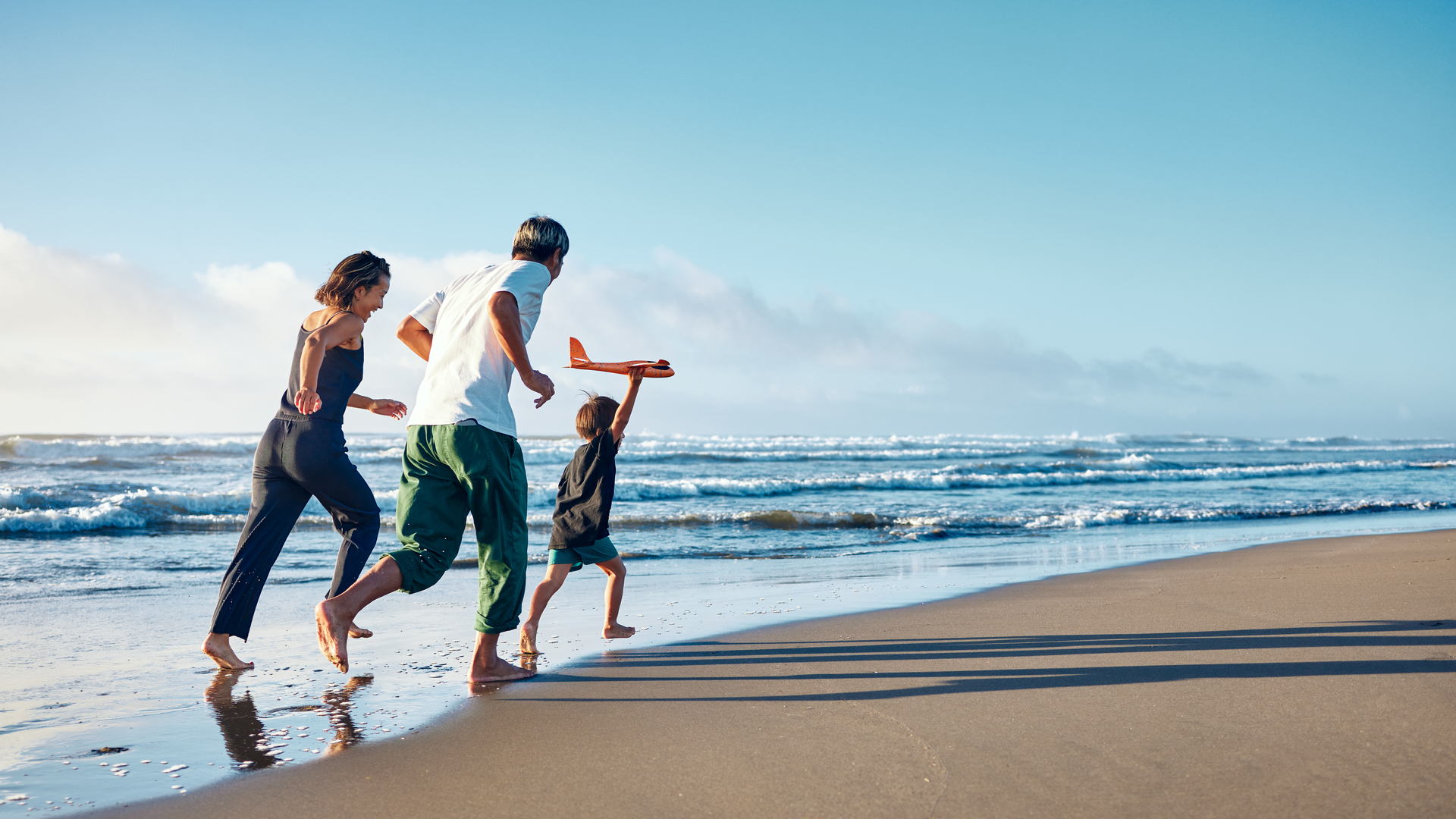 Parents running behind a boy running straight ahead with a model airplane held high in one hand.