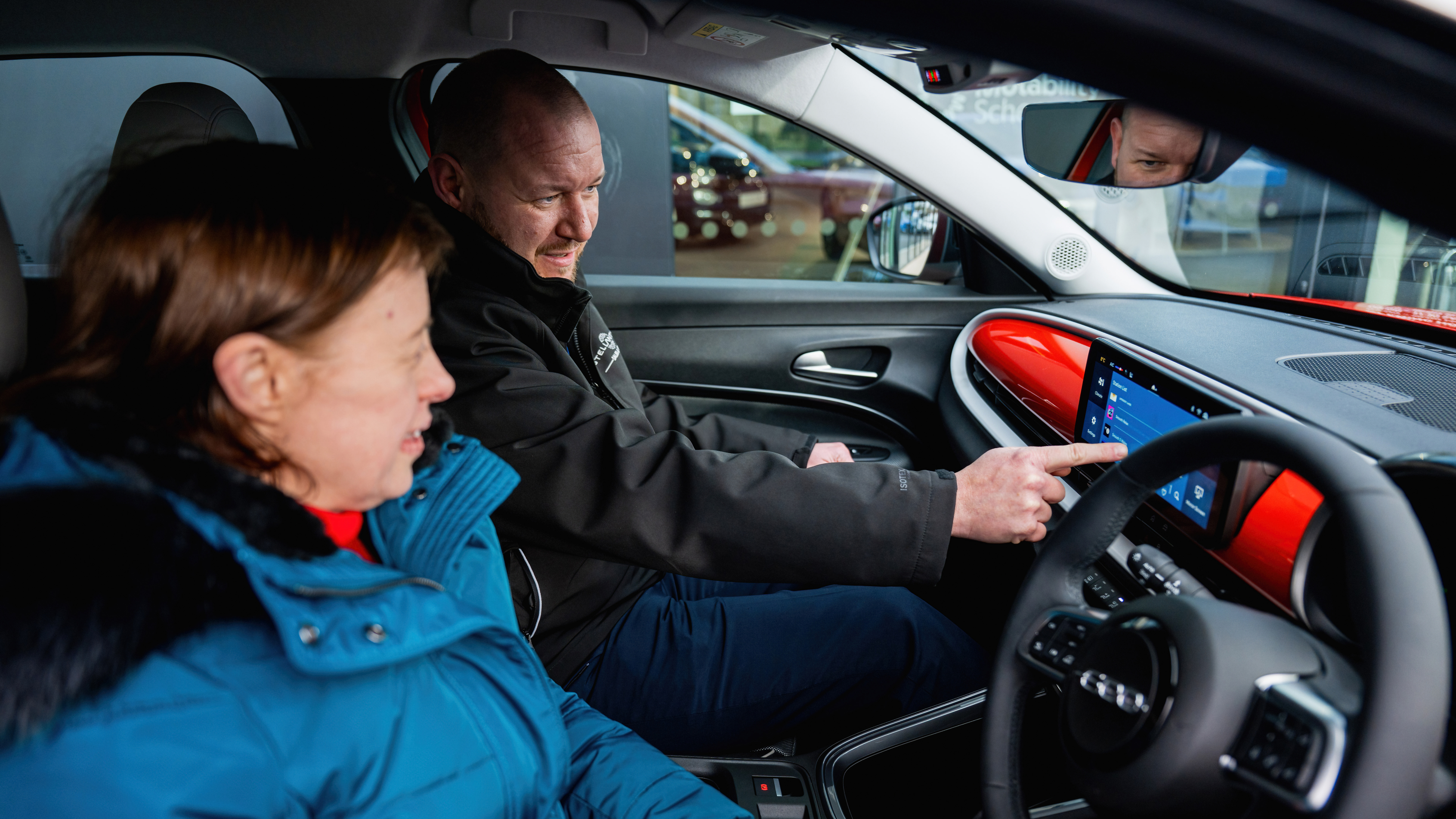 A man demonstrates a car’s touchscreen dashboard to a woman sitting in the passenger seat. Both are smiling as he explains the features, suggesting a friendly and informative vehicle handover or test drive experience.