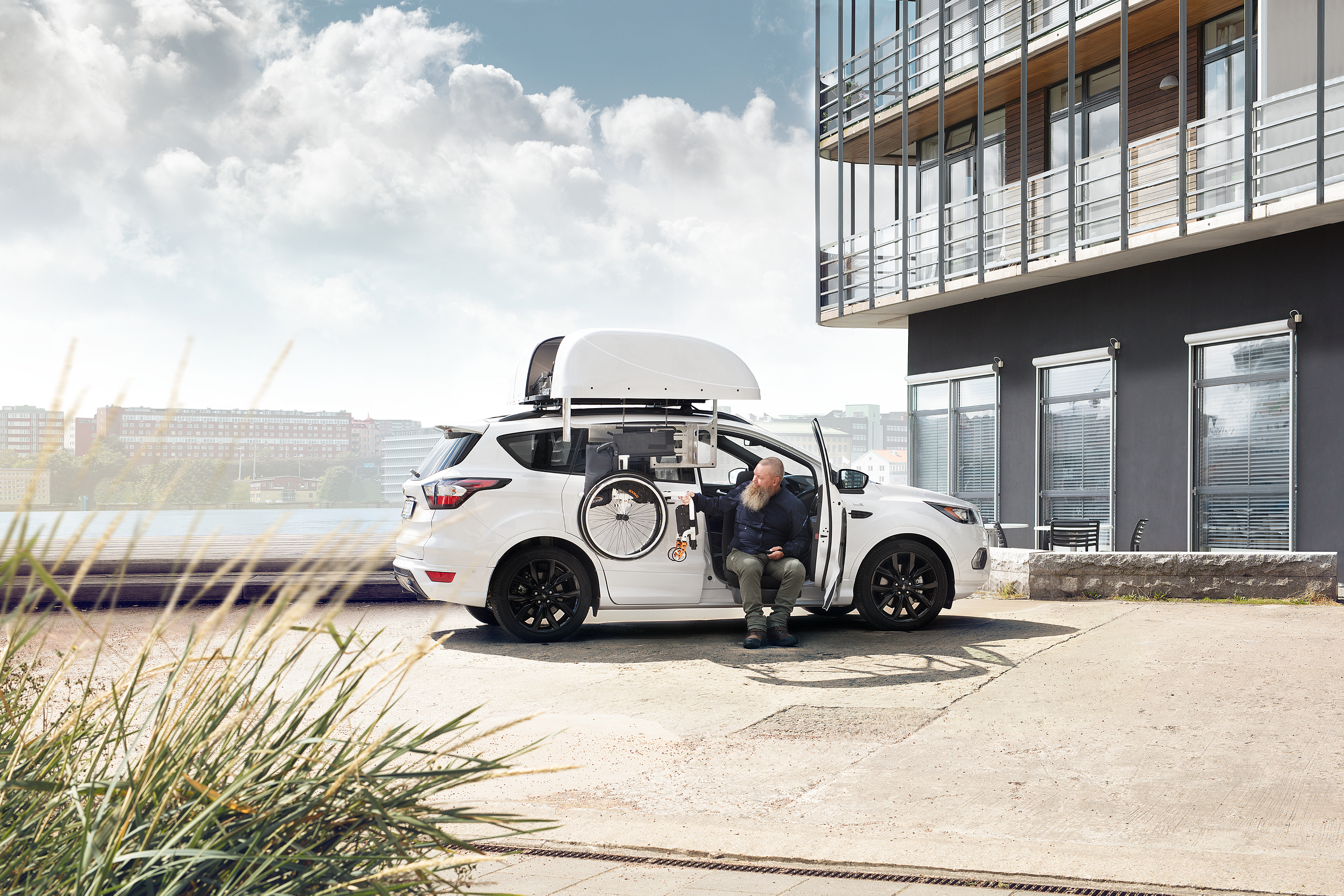 A man sits at the open side of a white SUV, preparing a mobility aid with a robotic arm that stores a wheelchair in a roof box. The vehicle is parked outside a modern building on a bright day, demonstrating advanced mobility equipment in use.