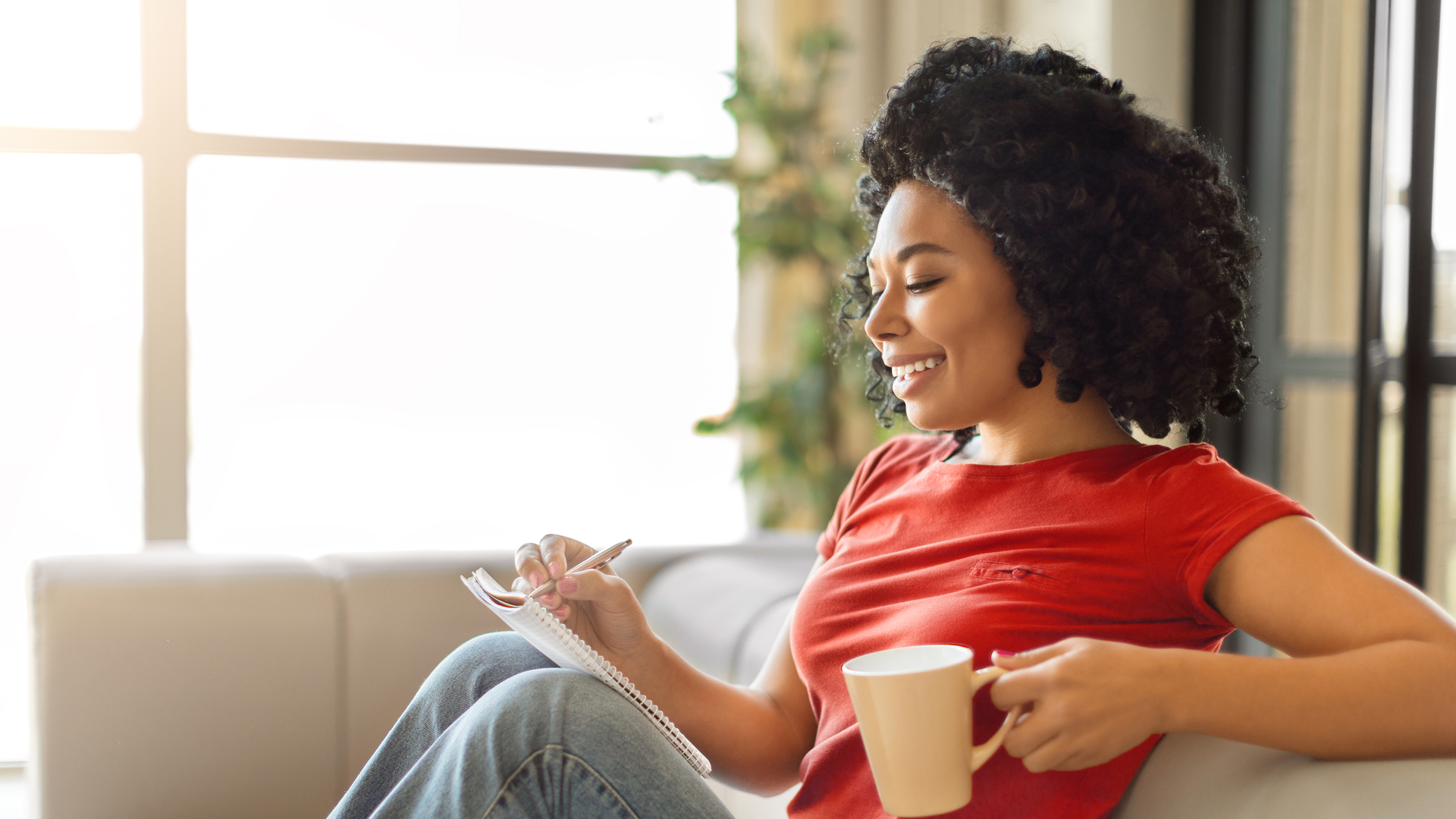 Smiling young black woman writing in notepad and holding mug of coffee