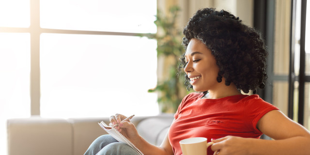 Smiling young black woman writing in notepad and holding mug of coffee