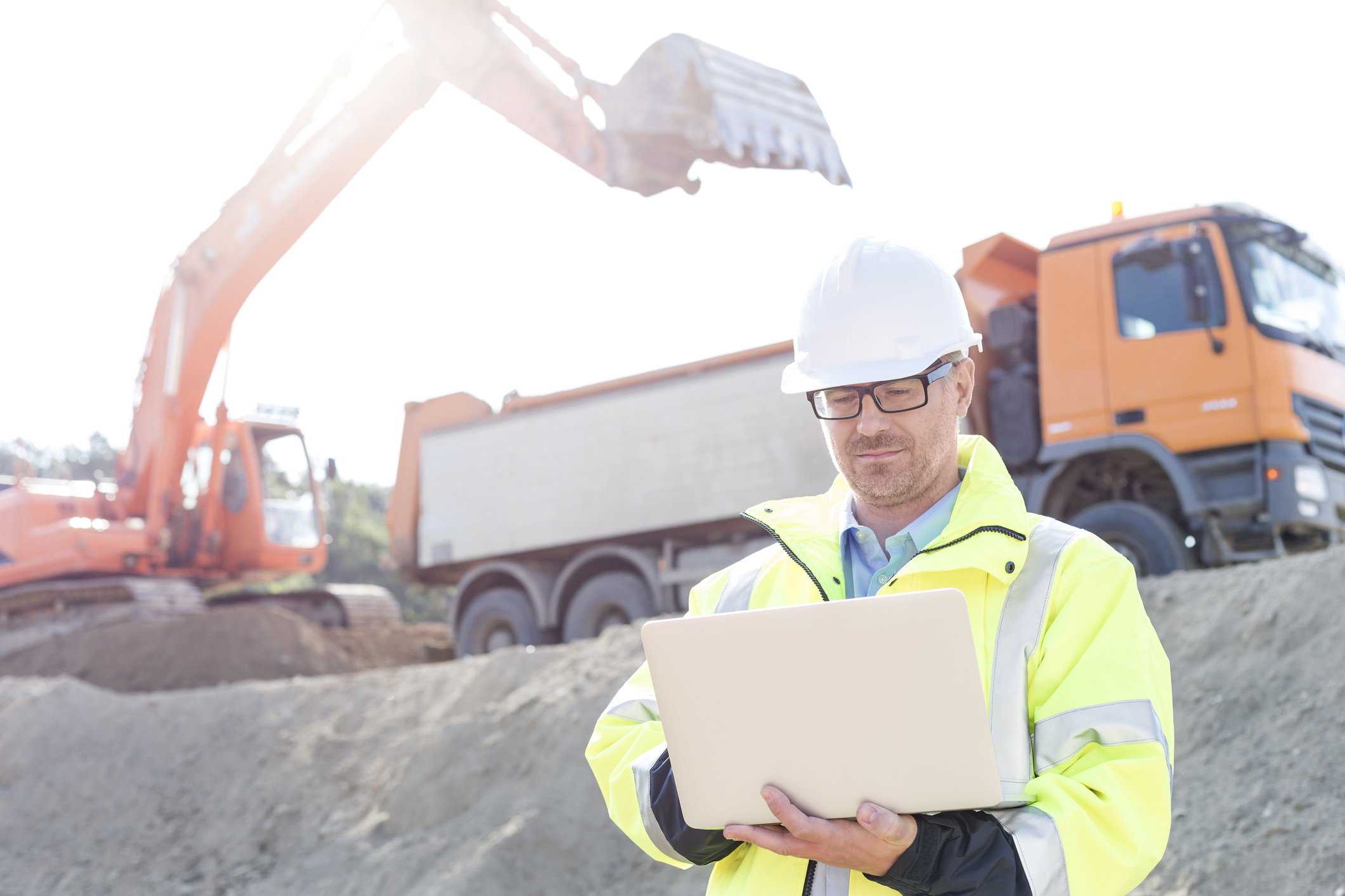 Man in hard hat and safety gear holding laptop with trucks and bulldozer in the background