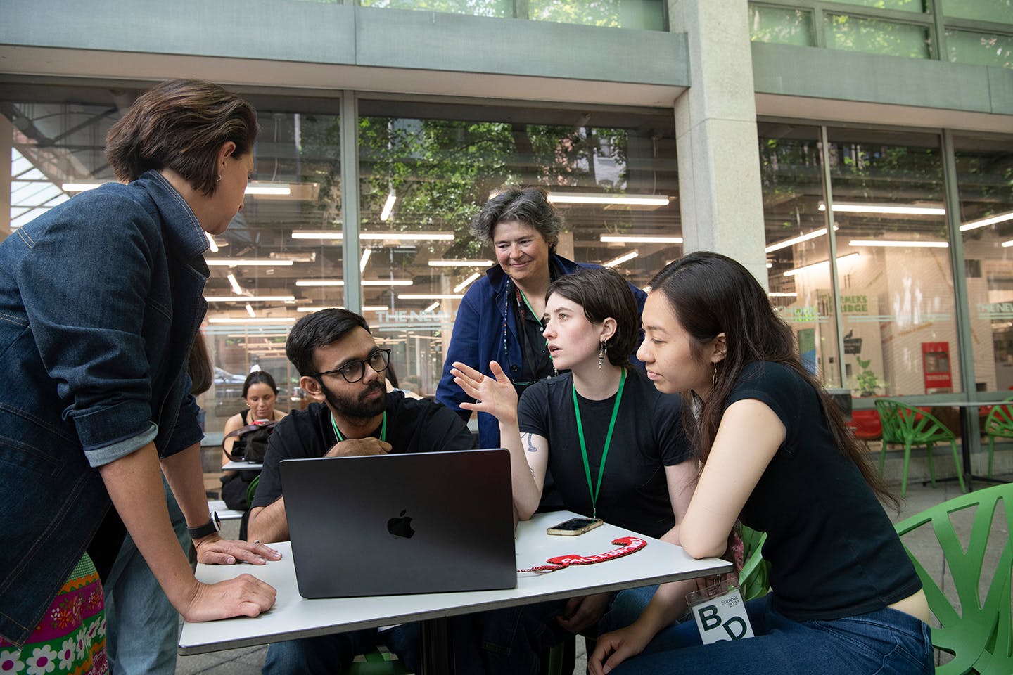 Two women stand over and observe three students, seated in an outdoor courtyard around a laptop, as they work.