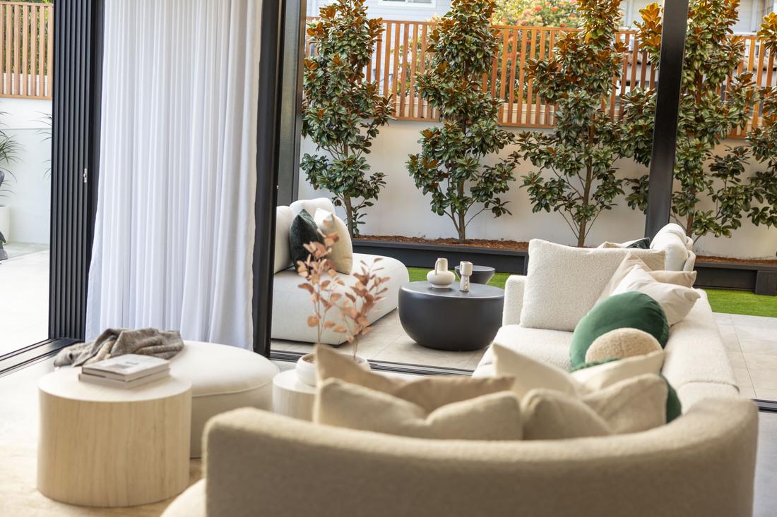Close up looking over a cream modular lounge through to the covered alfresco area with white couches. There are multiple tan and green cushions on the couch and a round coffee table and covered foot stool.