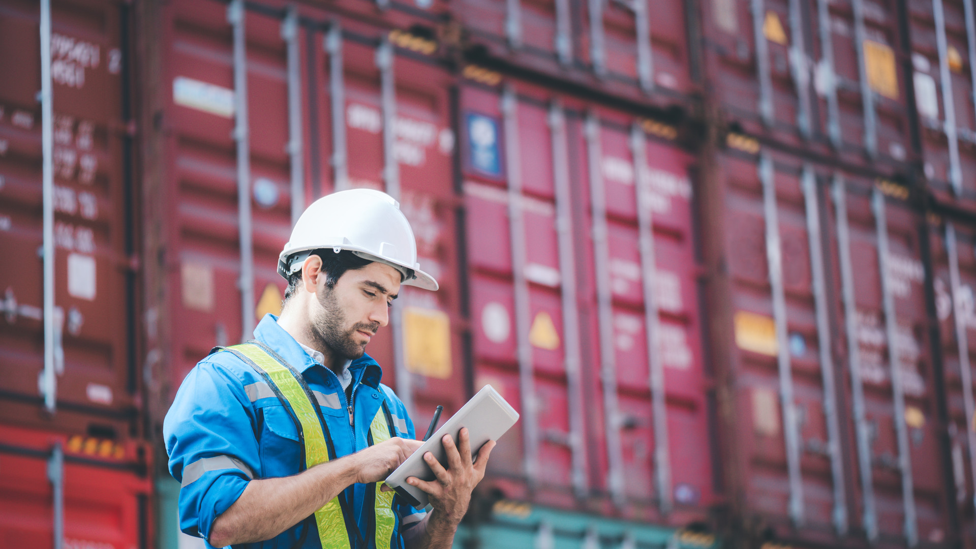 Man wears hardhat and reflection shirt and checking tablet with blurry metal containers in background. Concept of inventory and logistic management.