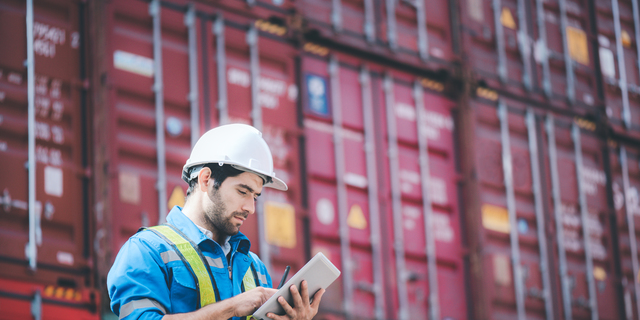 Man wears hardhat and reflection shirt and checking tablet with blurry metal containers in background. Concept of inventory and logistic management.