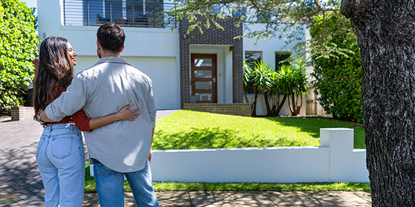 A man and a woman stand smiling with their arms around each other looking at a house