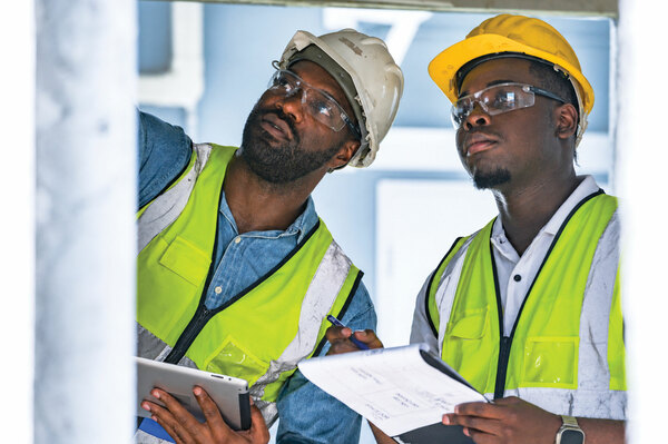 Two telecommunications workers wearing hard hats, safety glasses, and reflective vests review construction plans and inspect an installation site.