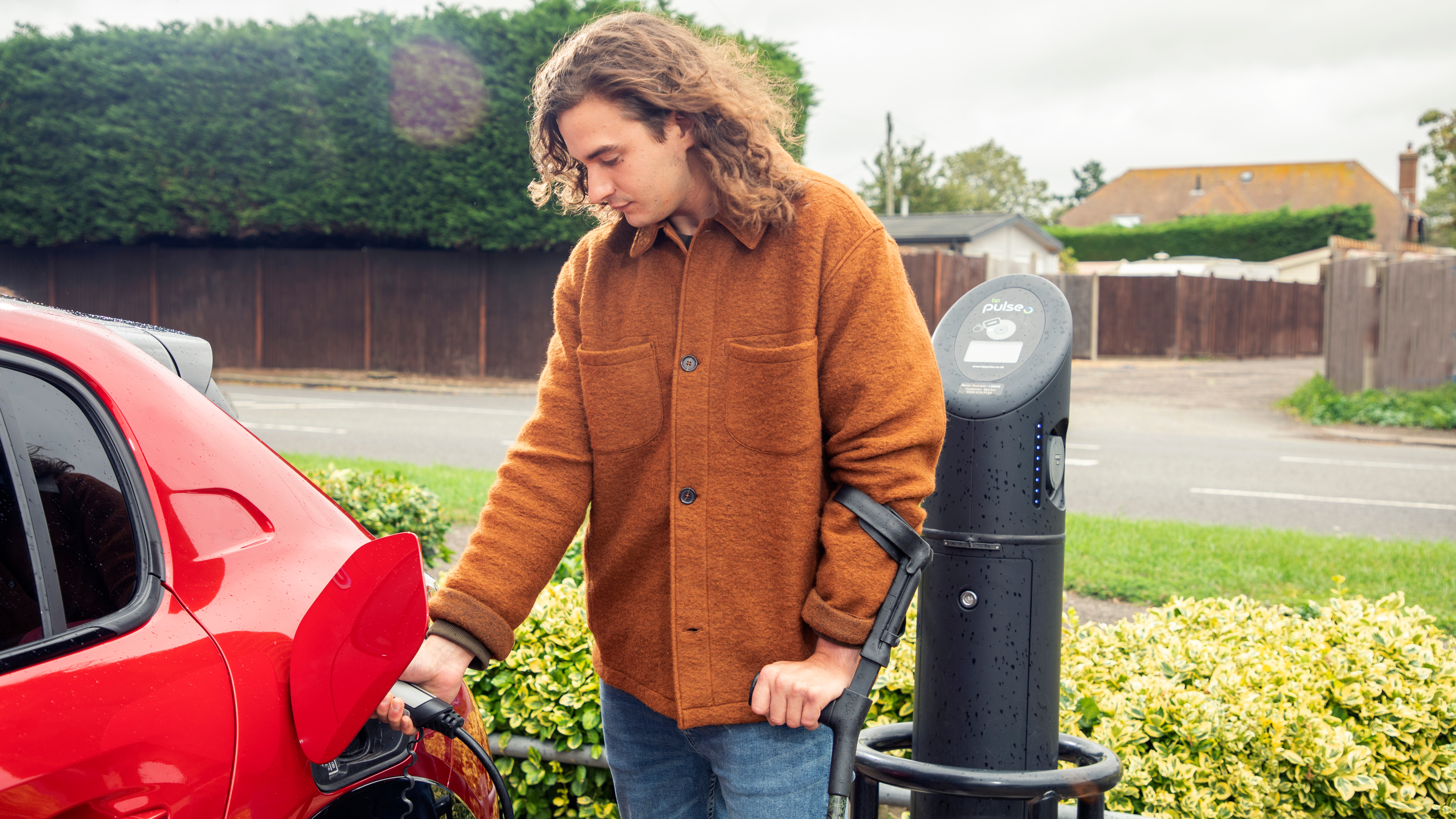 A man in a rust-coloured jacket charges a red electric car at a roadside EV charging point, holding a walking aid under one arm.