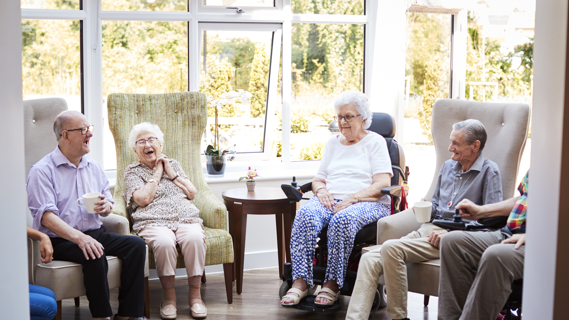 Male And Female Residents Sitting In Chairs And Talking In Lounge Of Retirement Home