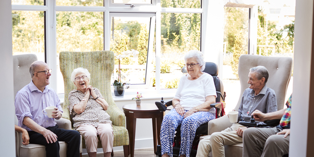 Male And Female Residents Sitting In Chairs And Talking In Lounge Of Retirement Home