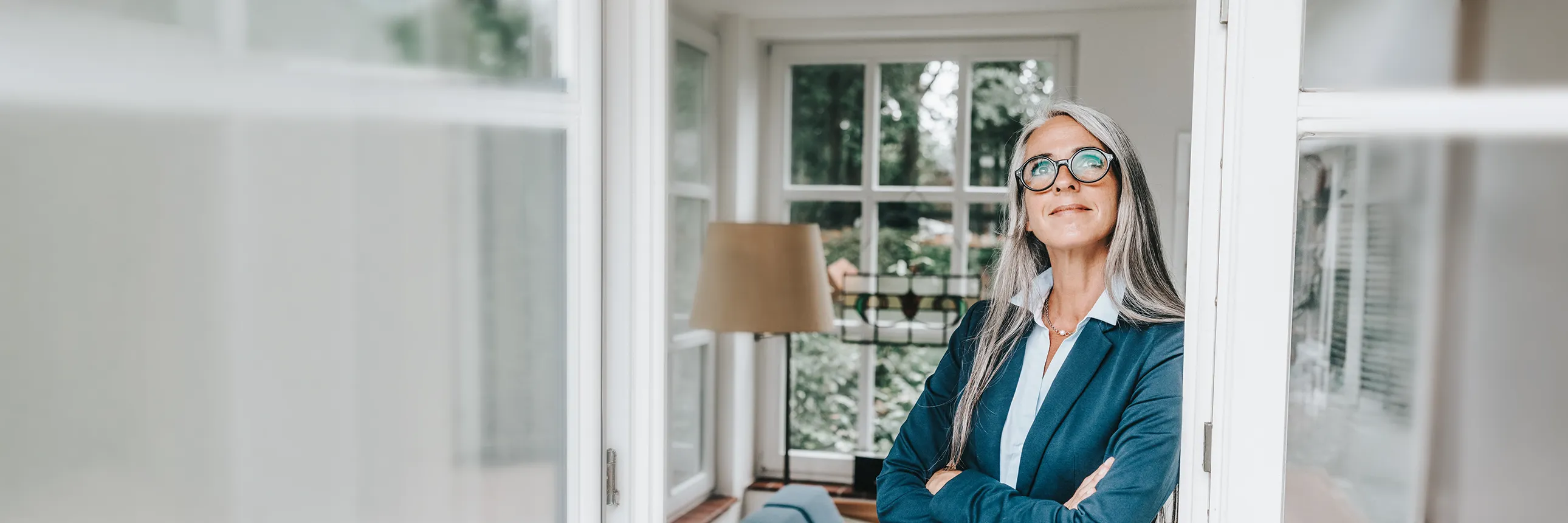 Person in business attire standing inside a home with arms crossed near large windows.