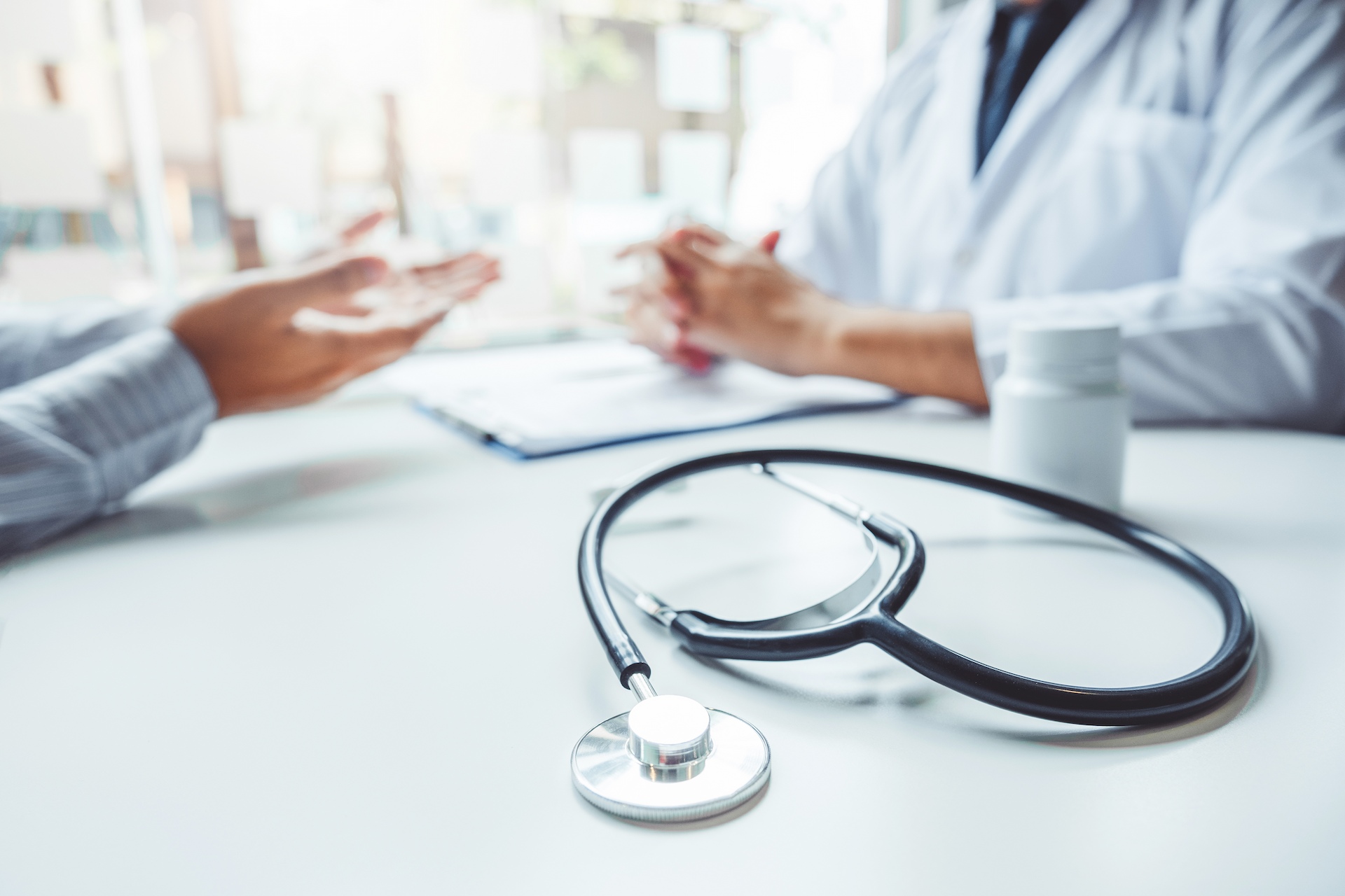 Photograph of a stethoscope in the foreground with a healthcare consultation taking place in the background.