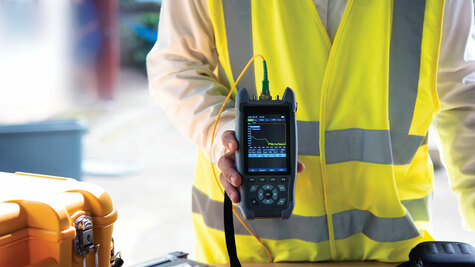 Technician in a high-visibility safety vest holding a handheld fiber optic OTDR testing device connected to a yellow cable