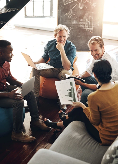 Four colleagues collaborating in a casual office setting, using laptops and reviewing a printed chart with green bar graphs.
