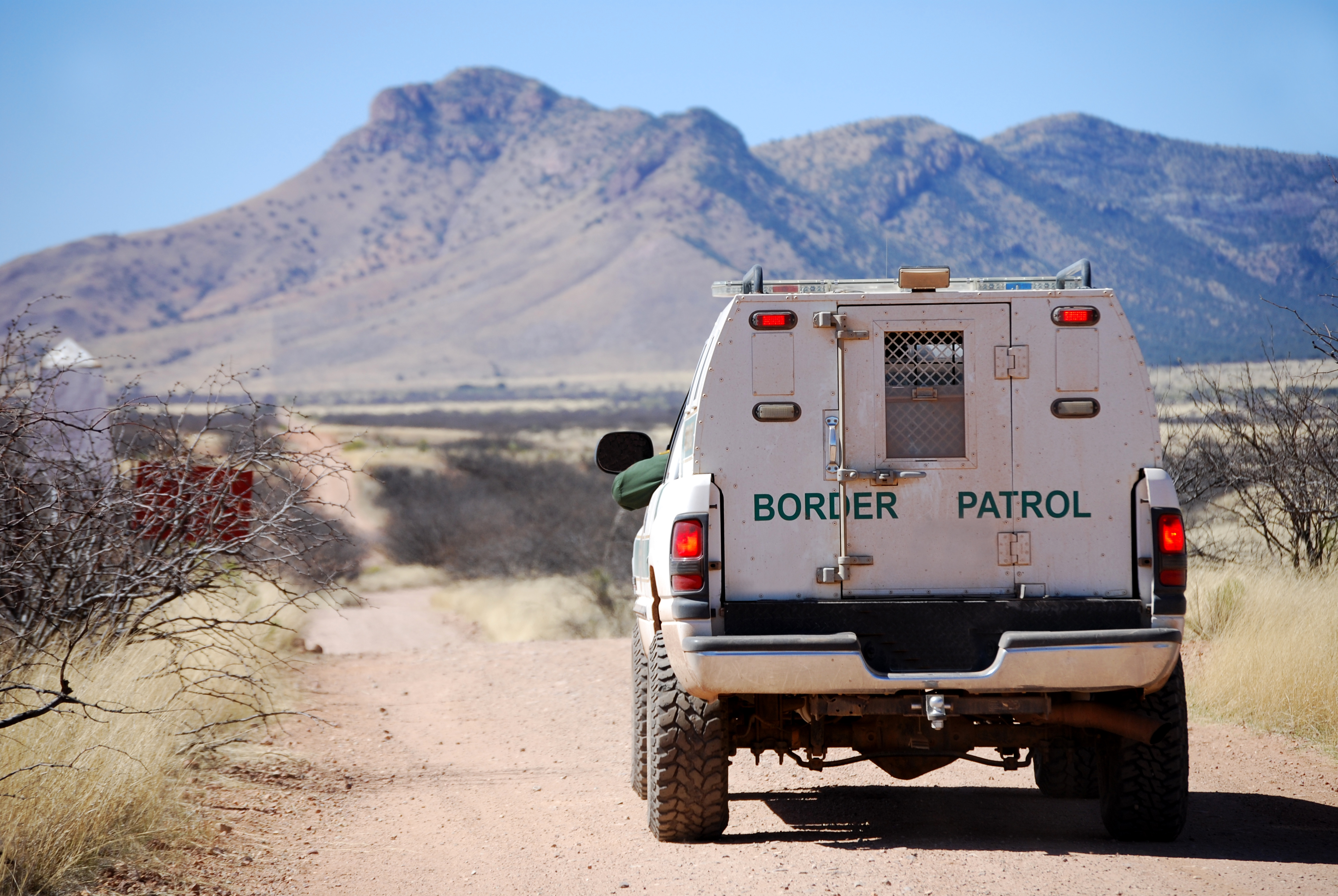A Homeland Security CBP vehicle drives down a dirt road in the desert towards a mountain.