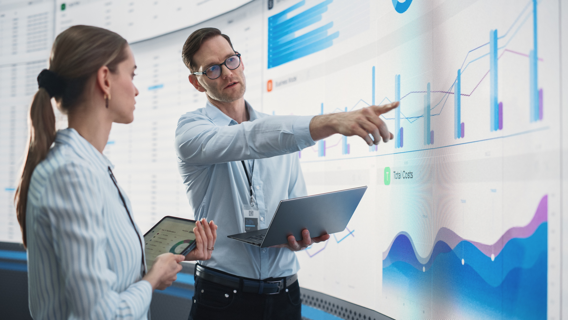 Caucasian Male And Female Data Scientists With Laptop And Tablet Standing Next To Big Digital Screen With Graphs And Charts In Monitoring Office. Colleagues Discussing Business Opportunities Or Risks.