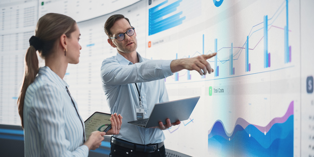 Caucasian Male And Female Data Scientists With Laptop And Tablet Standing Next To Big Digital Screen With Graphs And Charts In Monitoring Office. Colleagues Discussing Business Opportunities Or Risks.