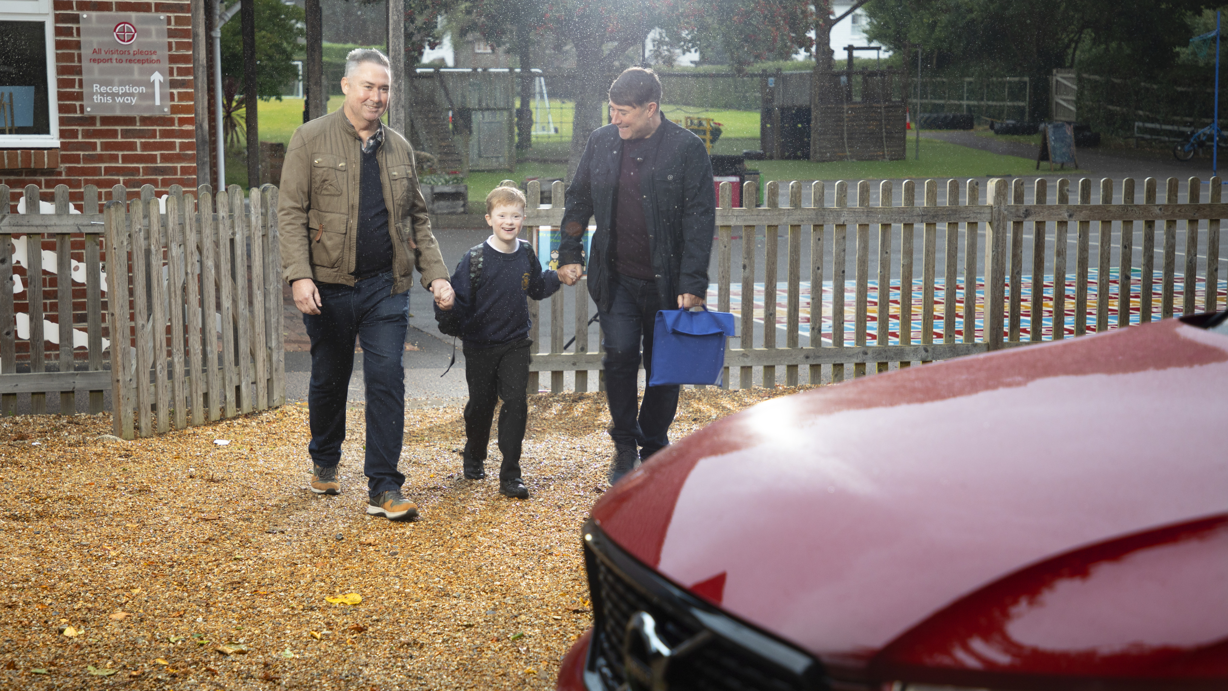 Two adults are walking a smiling young boy, likely just picked up from school, through a playground area on a rainy day. The boy is wearing a school uniform and backpack, holding hands with both adults. One adult carries a blue bag, and the group walks toward a red car in the foreground. A wooden fence and a school sign directing visitors to reception are visible in the background.