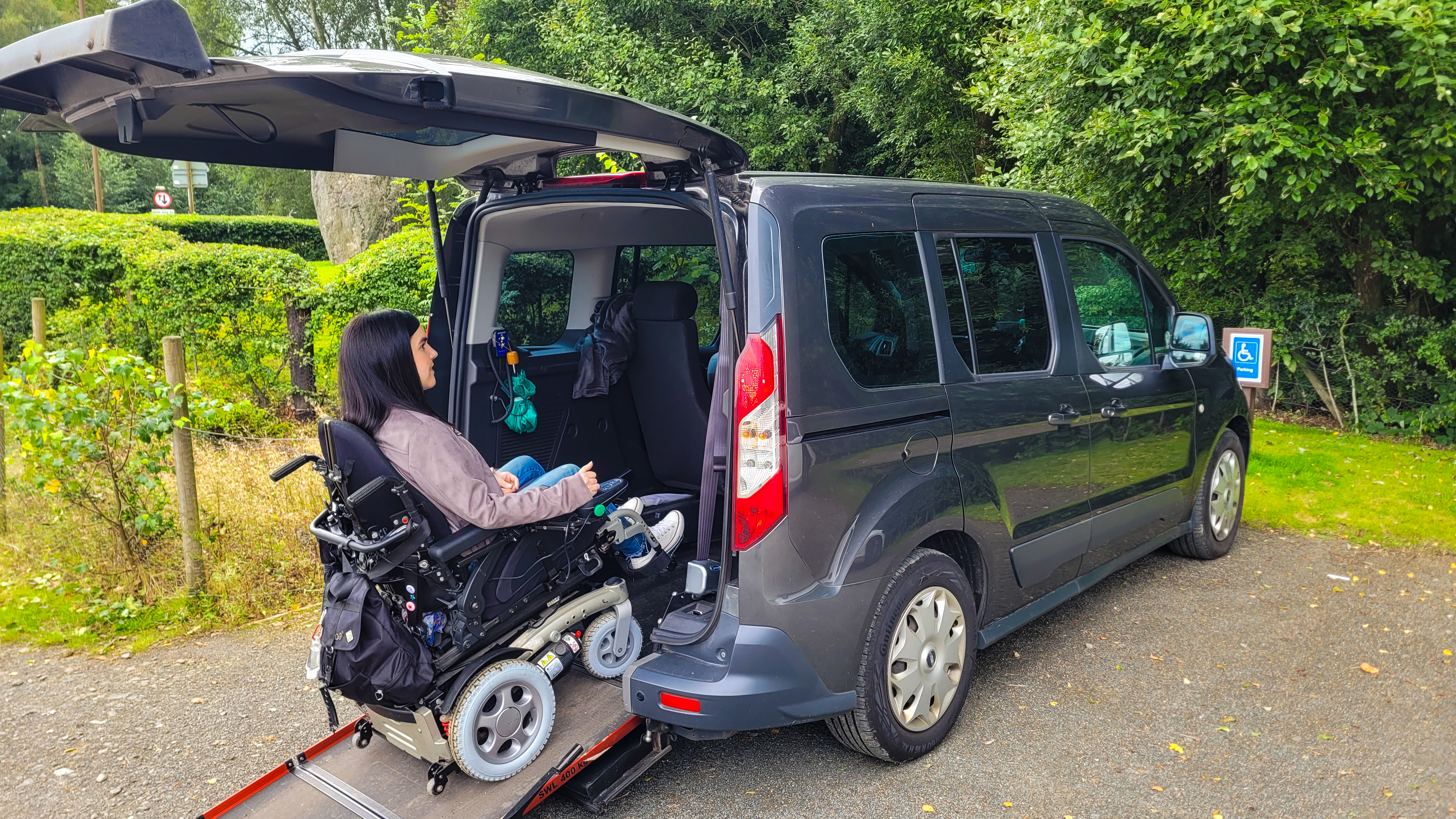 Emma Muldoon rides her powered wheelchair up the rear access ramp into a parked WAV in a designated accessible parking space, surrounded by greenery.