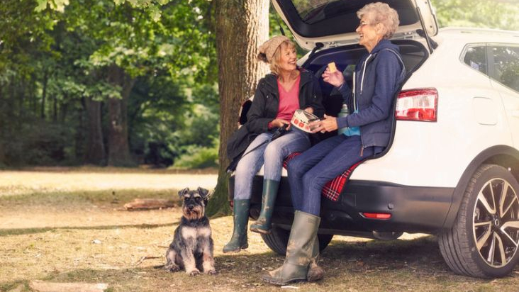 Two older women sit on the open boot of a white SUV, chatting and enjoying a snack in a wooded area. They are dressed in outdoor clothing with wellies, and a small grey and black dog sits attentively nearby. Sunlight filters through the trees, creating a warm and relaxed atmosphere
