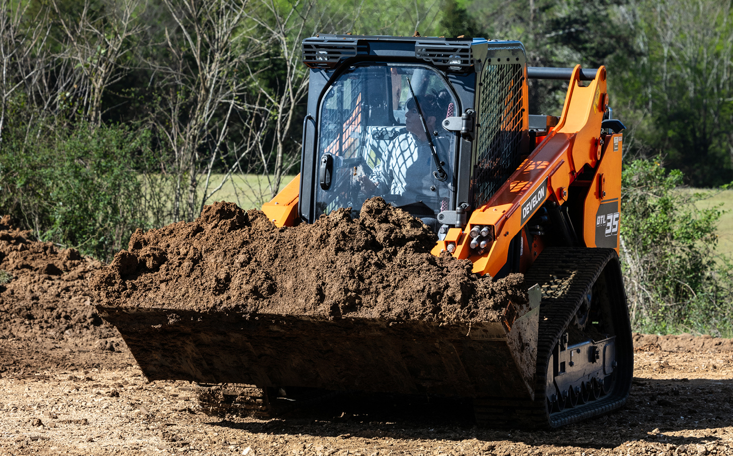 A DEVELON DTL35 compact track loader traveling with a bucket full of dirt on an open field.
