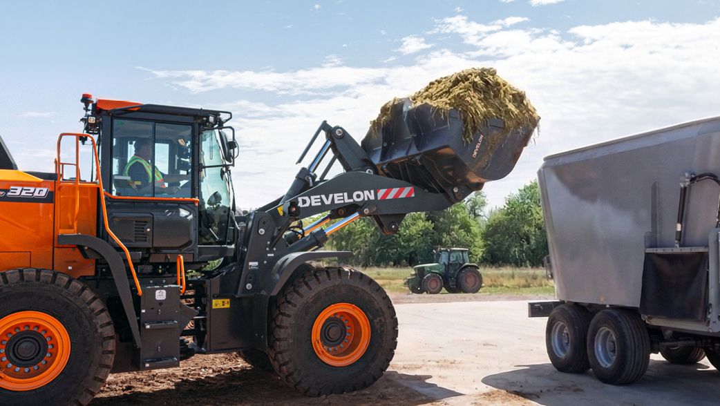 A DEVELON DL320-7 wheel loader dumping a load of hay into a trailer with a tractor in the background.