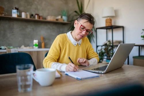Woman wearing red glasses and yellow sweater writing in a notebook while working on a laptop at home.